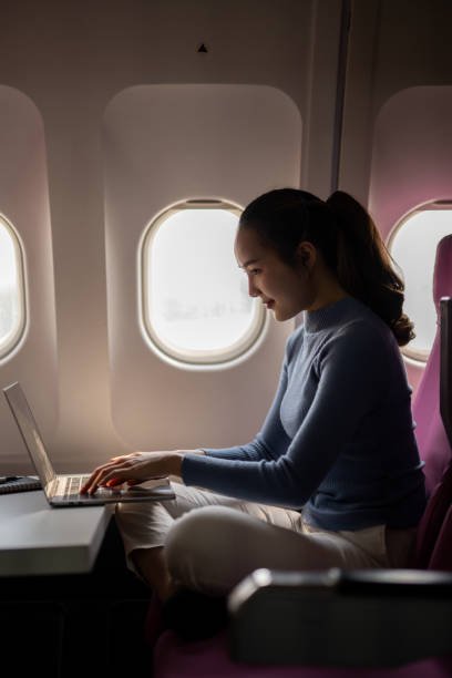 A young woman is using her laptop on a foldable tray table during a flight, with a window seat in an airplane cabin.